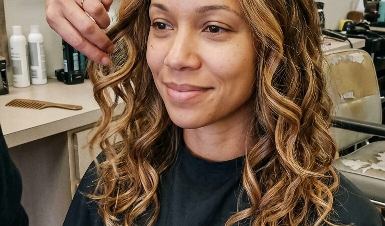 A smiling woman with medium-length warm brown hair and caramel blonde balayage highlights, styled in voluminous curls, sits in a salon with a stylist's hand touching her hair.