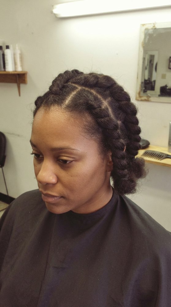 The hair is dark brown in color and appears to be thick and curly in texture. It is styled in two large cornrows that start at the crown and frame the face, with the ends curled and tucked under, creating a neat and elegant look. The hair seems to be of medium length, reaching just below the nape of the neck