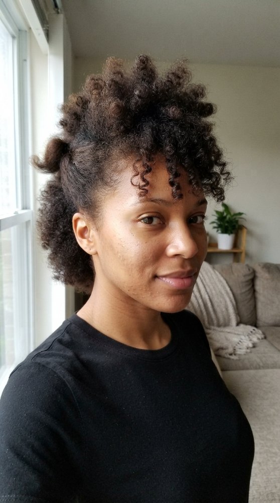 The hair is dark brown with a curly texture and appears to be at a medium length. It has a natural, voluminous look with tight curls that are more defined near the face and on top, gradually loosening towards the back and sides. There are dimensional color, and the hair does not have a specific cut style or layers, but rather a puffed, natural shape