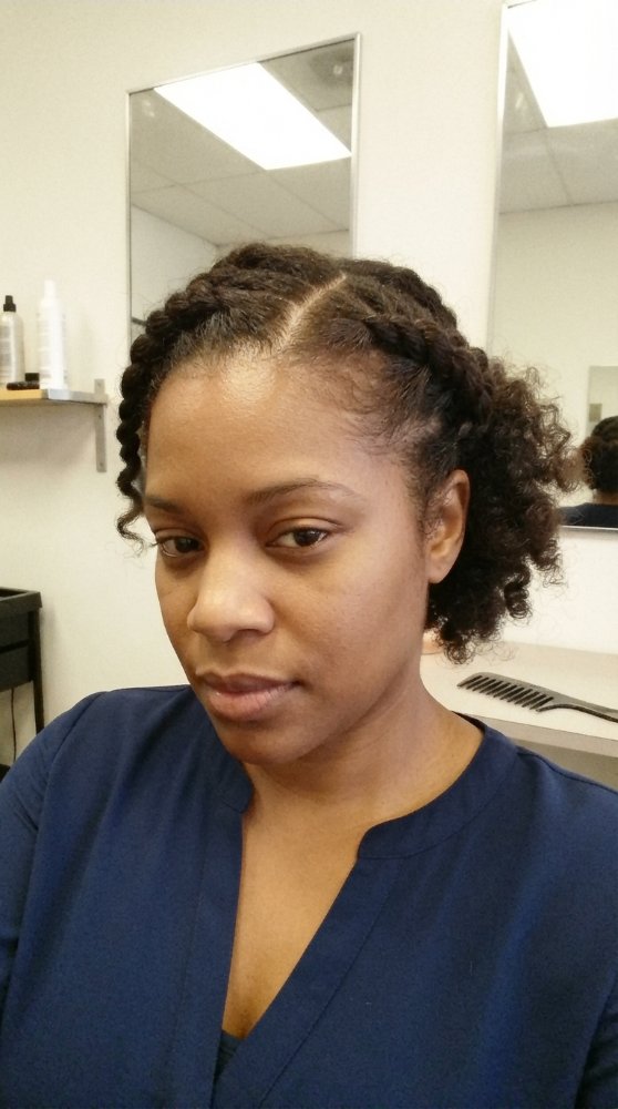 The hair is dark brown with a curly texture and appears to be styled in two braids that are pulled back and secured, with loose curls framing the face. The hair length seems to be around chin or shoulder-length, and there are dimensional color. The cut style is not clearly visible, but it appears to have some layers, and there is visible regrowth or gray blending