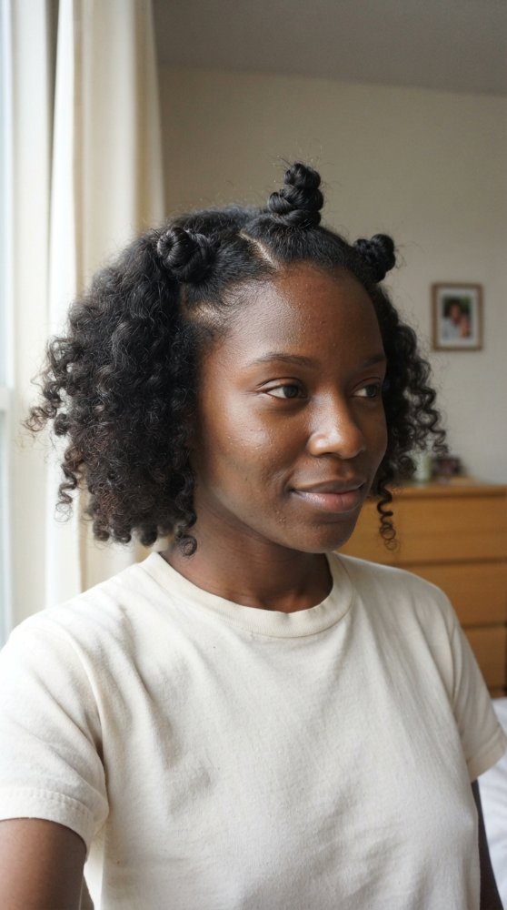 The hair is dark brown with a curly texture and medium length. It is styled in space buns on top of the head, with the rest of the hair left loose and falling around the face and shoulders in loose curls. The hair appears to have a natural, effortless look with dimensional color, and obvious cut style or layers are visible, but it does have a face-framing effect