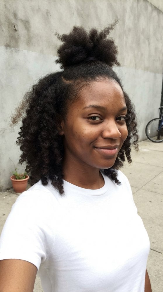 The hair is dark brown with a curly texture and medium length. It is styled in a half-up half-down look, with the top section pulled into a puff and the bottom section left loose in tight curls that frame the face. The hair appears to have a natural, effortless look with dimensional color, and there is evidence of braids or regrowth visible