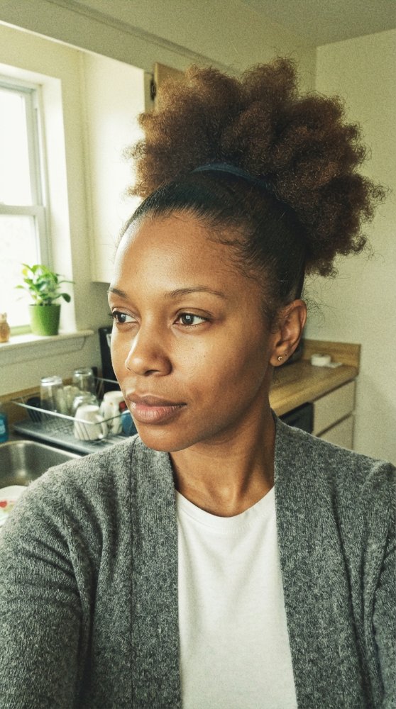 The hair is dark brown with a curly texture, pulled up into a high puff secured by a black hairband. The hair appears to be of medium length, with dimensional color, and discernible cut style, layers, curls, braids or face-framing elements. There is also visible dimension, gray blending or regrowth