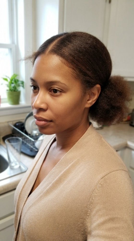 The hair is dark brown with a low ponytail or bun style, pulled back from the face. The texture appears to be thick and curly, with a natural, effortless look. There are dimensional color, and the hair seems to be in its natural state with braids or curls, but rather a smooth, sleek finish on the top section and a puffier texture at the back