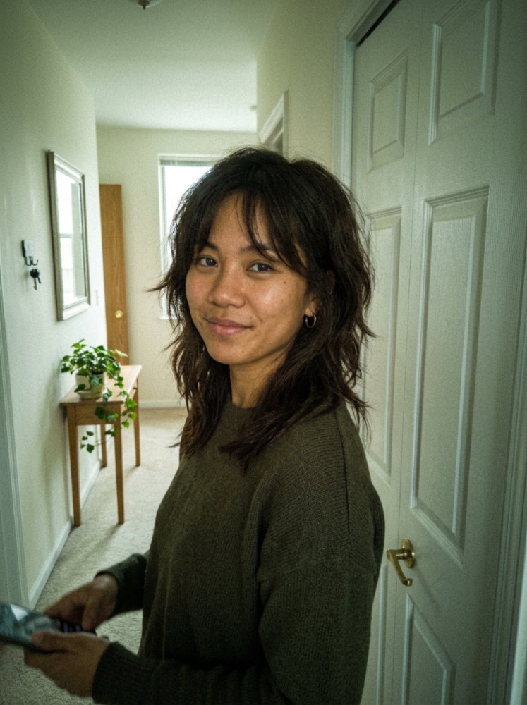 The hair is dark brown with a medium length and a wavy texture. It features layers that frame the face, with choppy ends giving it a piecey look, and appears to have subtle dimension with dimensional color. There are braids or curls, and visible regrowth or gray blending