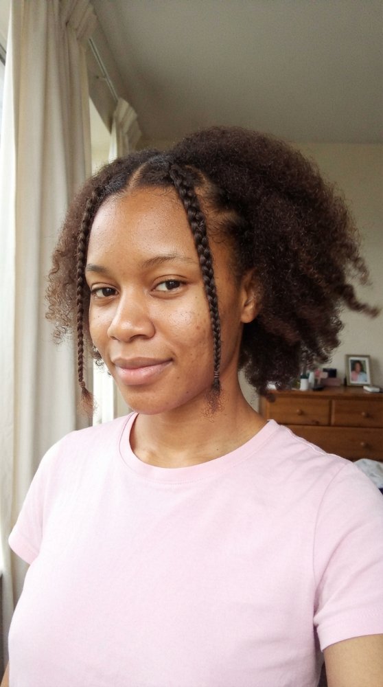 The hair is dark brown with a natural, curly texture and appears to be medium in length. Two braids frame the face, while the rest of the hair is loose and puffy, showcasing its natural curl pattern. There is visible evidence of highlights, lowlights, balayage, toner, gloss or gray blending, and the cut style seems to be a simple, natural shape with obvious layers or face-framing elements