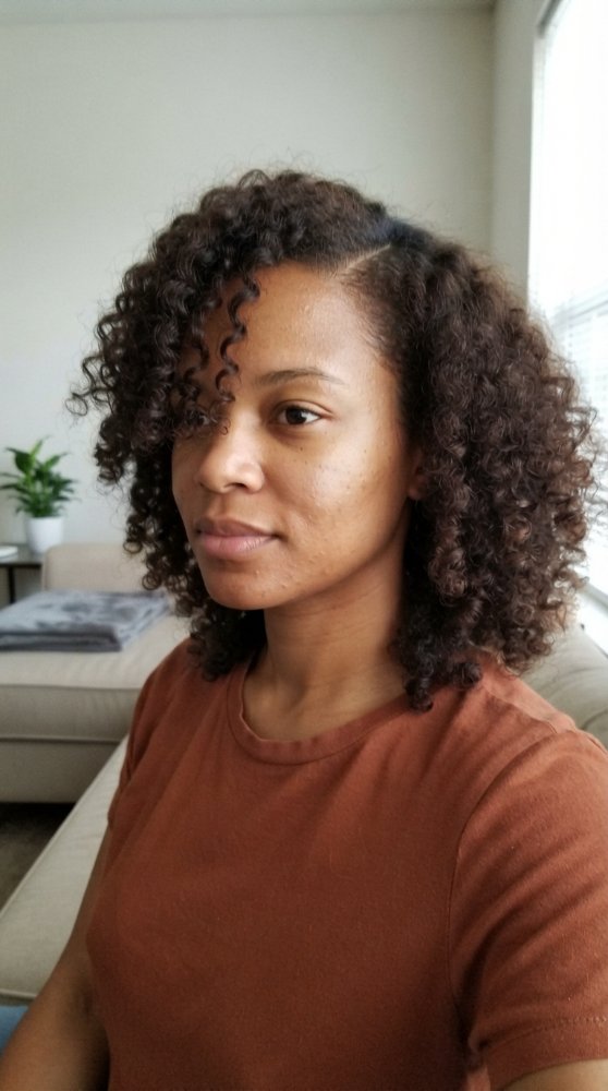 The hair is dark brown with a rich, deep color and a curly texture. It falls just above the shoulders in a shoulder-length cut with layers that enhance the natural curl pattern, which is tight and bouncy with a lot of movement. The curls frame the face and have a lot of dimension, but there are visible highlights, lowlights, balayage, toner, gloss, braids or gray blending