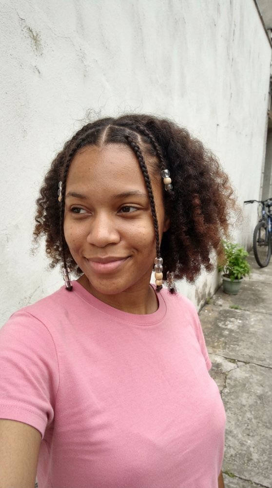 The hair is dark brown with subtle hints of lighter brown and reddish tones, particularly at the ends. It is styled in a short, curly cut with braids framing the face, and the braids are adorned with beads. The texture appears to be tightly coiled and curly, with a natural, effortless look