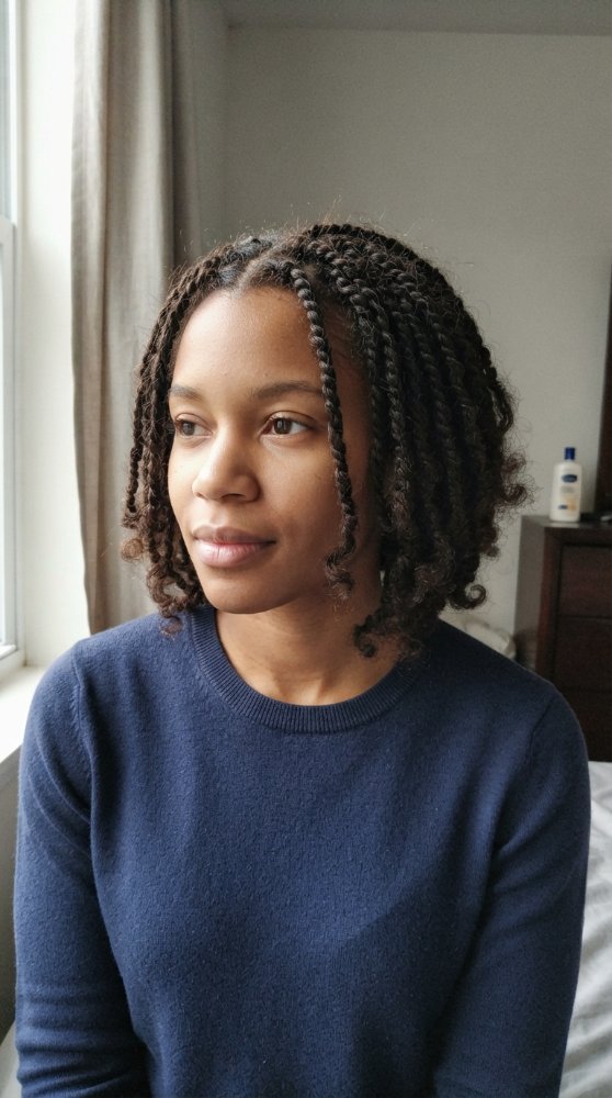 The hair is dark brown with a subtle sheen, styled in small, tight braids that fall just below the chin in length. The texture appears to be thick and coarse, with a defined curl pattern visible at the ends of the braids, which have a slightly looser curl. There is visible evidence of dimensional color, and the cut style is uniform with discernible layers or face-framing elements. The braids are neatly done with regrowth or gray blending visible