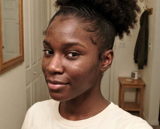 The hair is dark brown and tightly coiled, with a high ponytail or puff style that adds volume. The texture appears to be curly and kinky, with a defined edge around the face and a smooth, sleek finish on the sides, suggesting braids or cornrows. The hair has dimension with the puff on top adding height