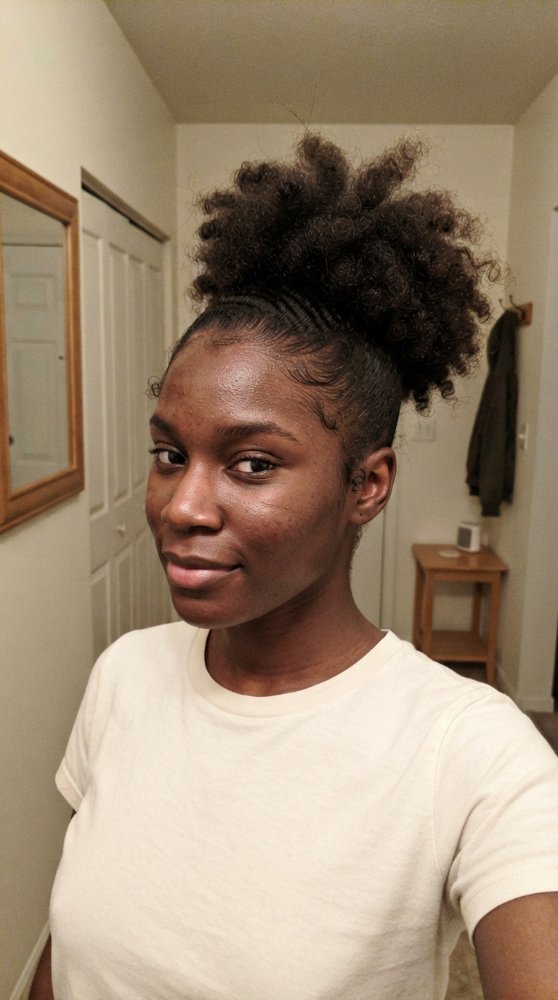 The hair is dark brown and tightly coiled, with a high ponytail or puff style that adds volume. The texture appears to be curly and kinky, with a defined edge around the face and a smooth, sleek finish on the sides, suggesting braids or cornrows. The hair has dimension with the puff on top adding height