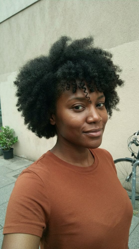 The hair visible is dark brown with a natural texture that appears to be tightly coiled and curly. It is cut in a style that frames the face with a voluminous, rounded shape that reaches the chin or the collarbone in length. The curls are tight and bouncy, with some looser, face-framing tendrils around the forehead. There is visible evidence of highlights, lowlights, balayage, toner, gloss, braids or gray blending