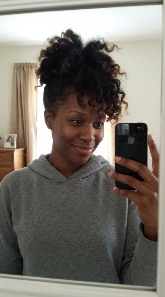 The hair visible in this is dark brown with subtle hints of lighter brown, particularly at the ends. It appears to be tightly coiled and curly, pulled up into a high puff, showcasing its natural texture. The hair has dimension with dimensional color, and styling such as braids or face-framing layers