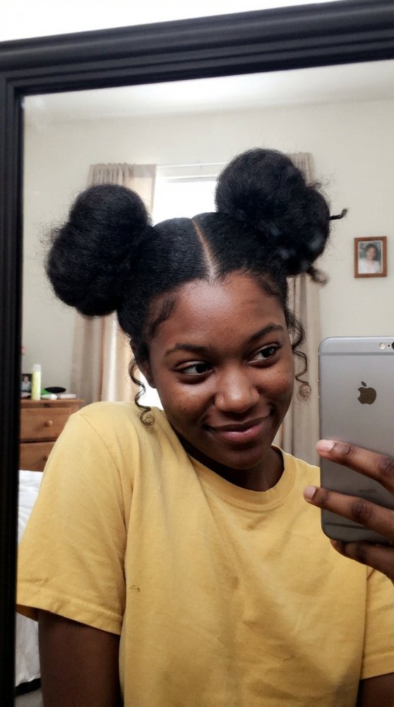 The hair visible in the is dark in color and appears to be tightly coiled and dense in texture. It is styled in two large buns on top of the head, with a center parting and some loose, curly tendrils framing the face. The hair has dimensional color, and its cut style is not discernible, but it has been sectioned and smoothed to create the buns, with layers or braids visible